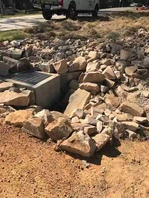 A concrete drainage headwall with a grate, ringed by rough rock riprap on a red-dirt slope