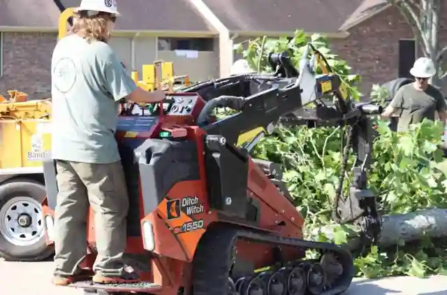 An Artmire's crew member in a roundel tee driving a Ditch Witch mini loader with a trunk on a brick-home street